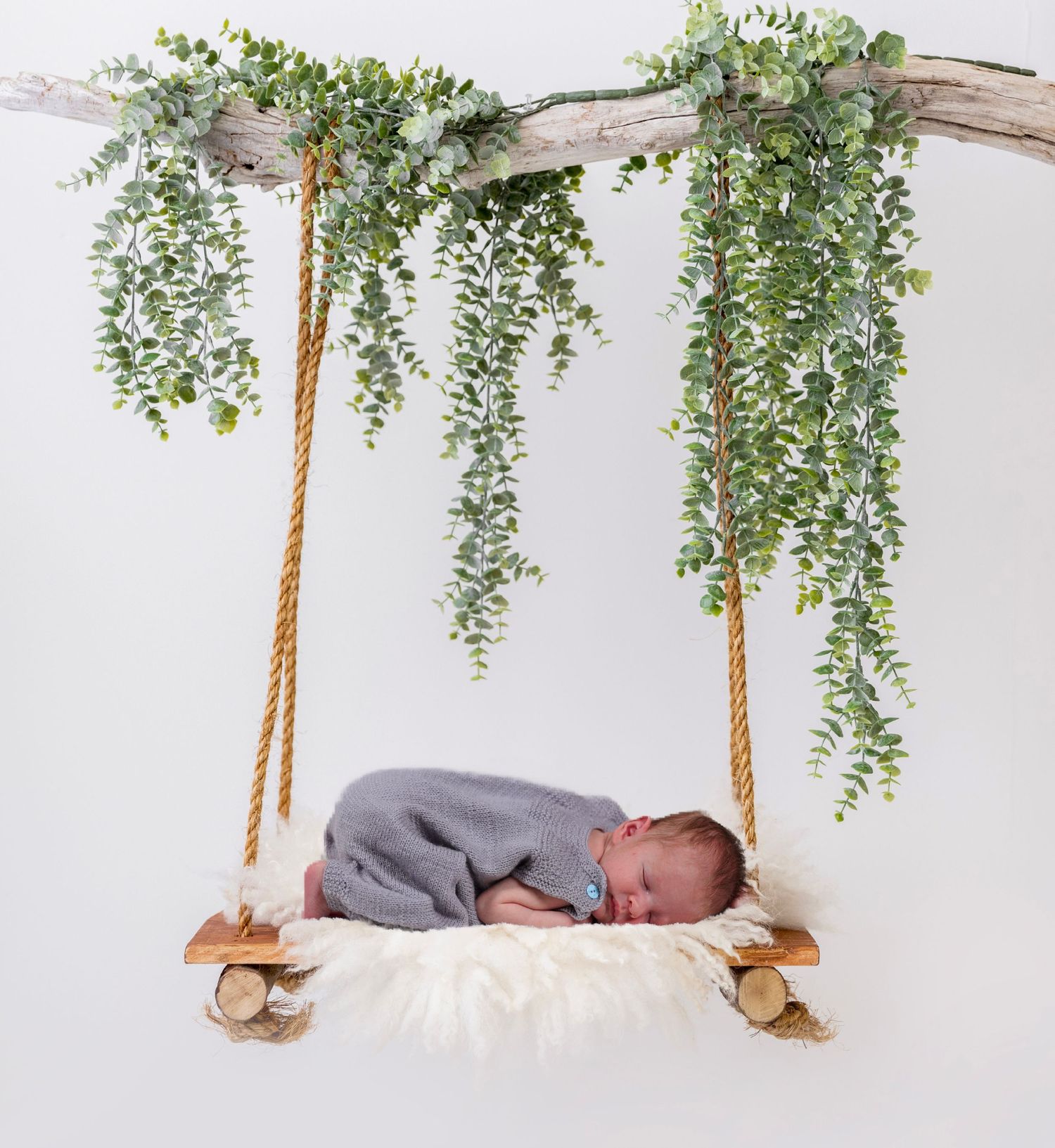 Sleeping newborn baby in grey wrap resting on a swing decorated with greenery.