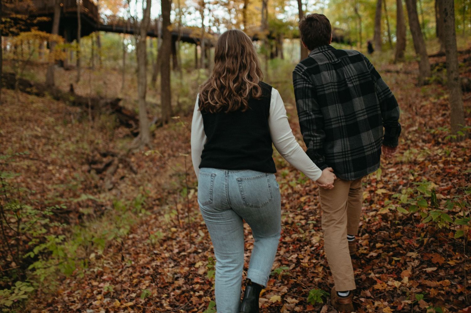 A couple walks hand in hand through a forest trail covered in fallen autumn leaves.