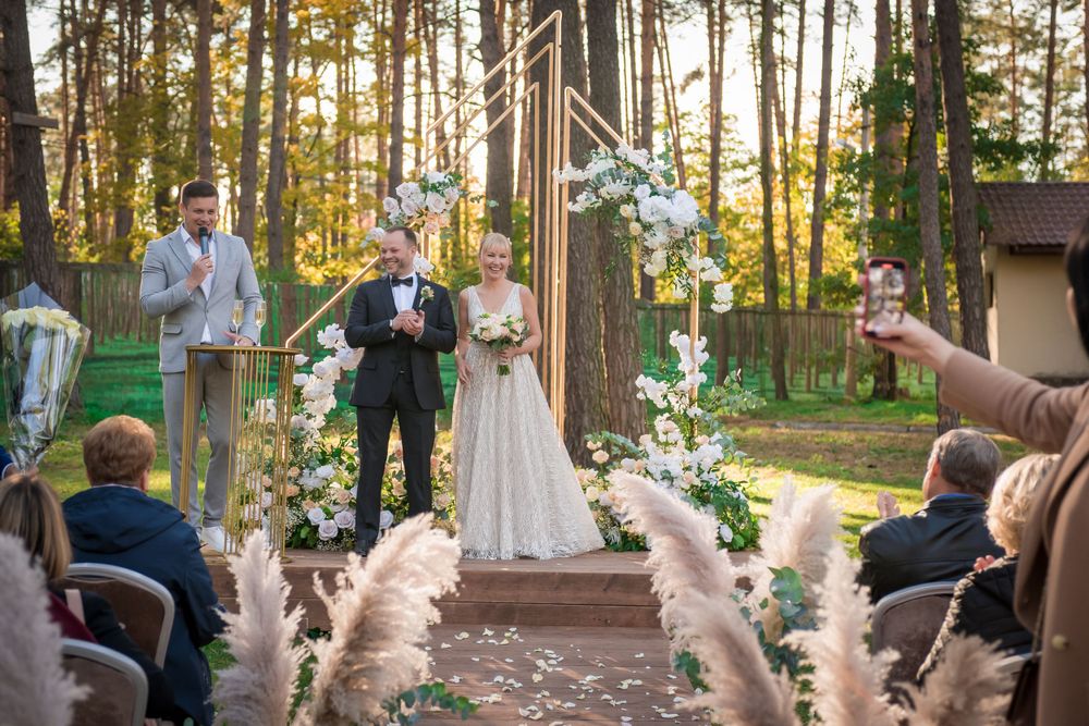 Wedding ceremony taking place in a sunlit forest setting with guests seated on white chairs.