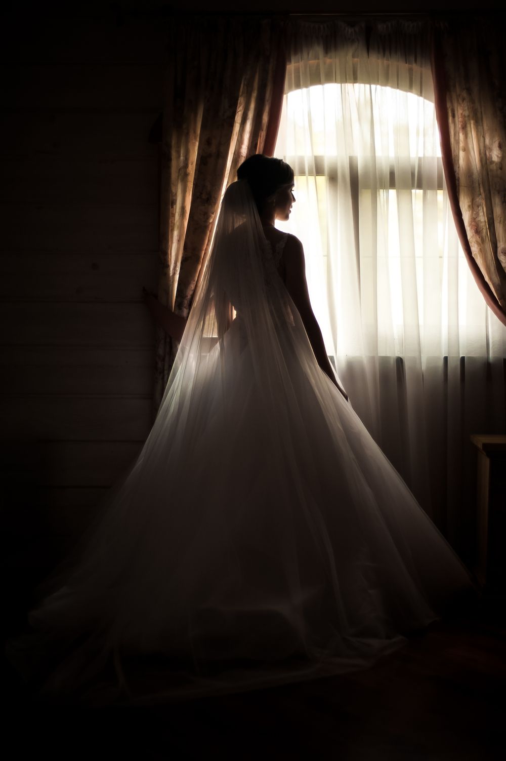 Silhouette of wedding dress against window light creating dramatic shadows.