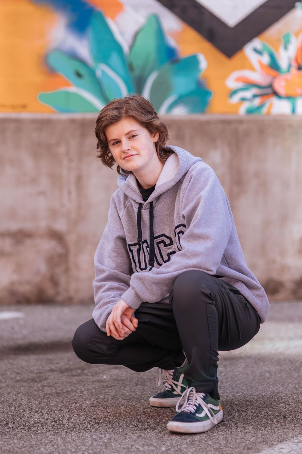 Student in gray UCD sweatshirt crouches in front of vibrant graffiti wall.