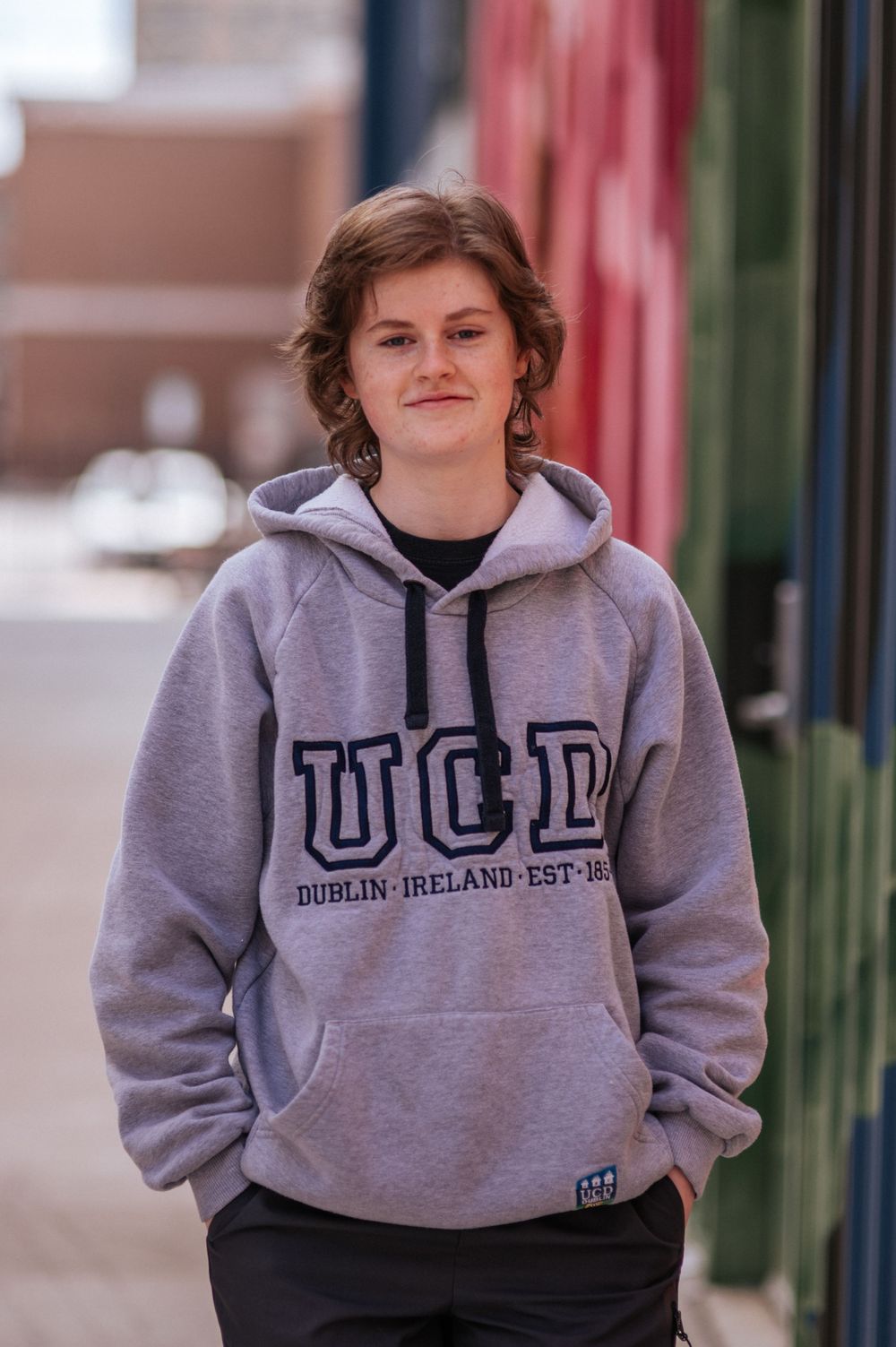 Student wearing gray UCD hoodie stands casually on city street.