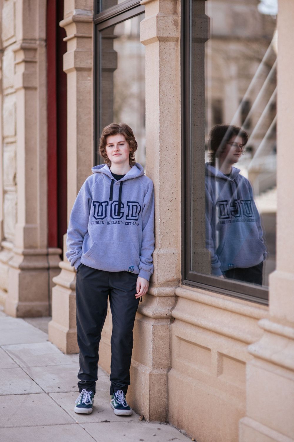 Student in UCD sweatshirt stands by reflective window with stone column architecture.