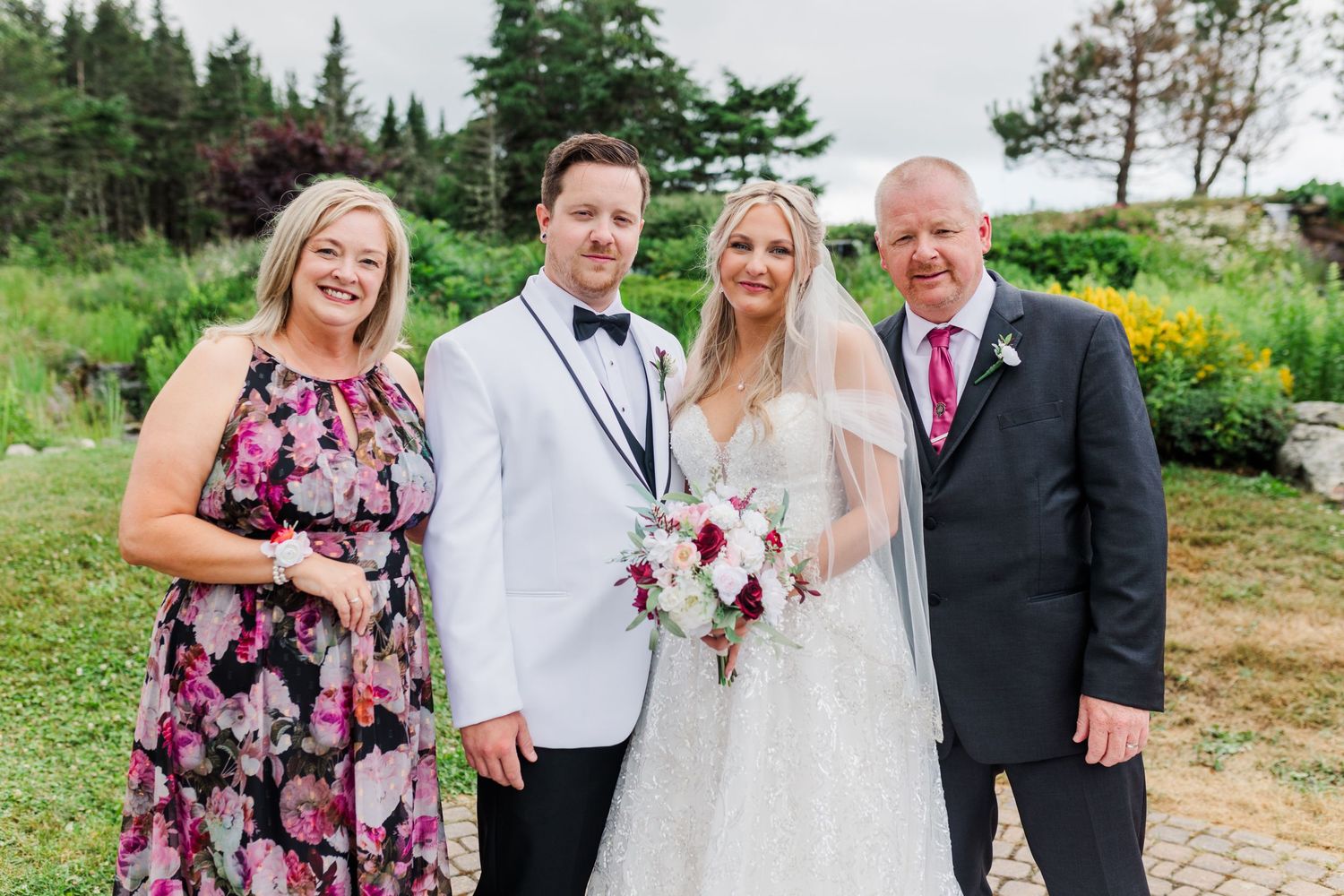 Wedding couple poses with family members in garden setting with evergreen trees in background.