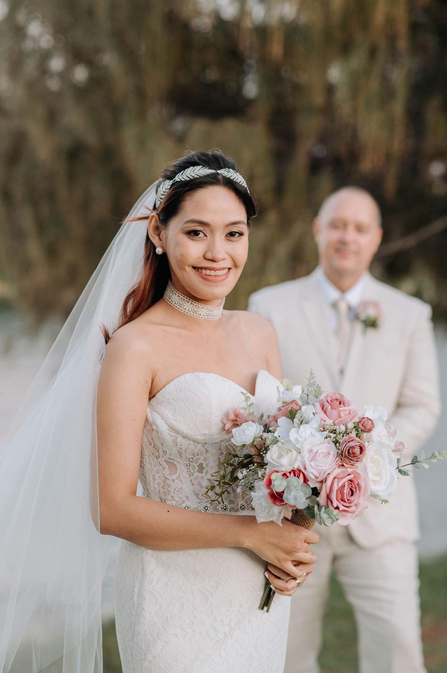 Elegant bride holds pink and white bouquet in lace wedding dress with veil outdoors.