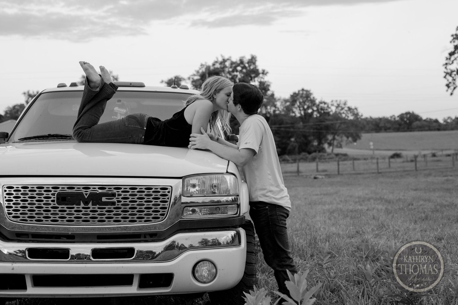 A black and white photo of a couple sharing a sweet moment against their truck in a rural setting.