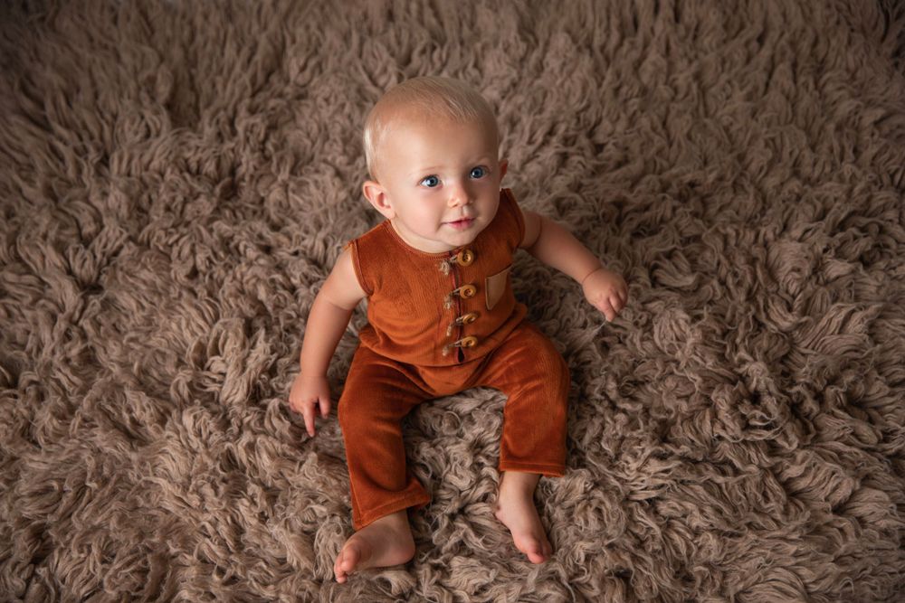 Cute baby in rust orange romper sitting on fluffy brown textured rug.