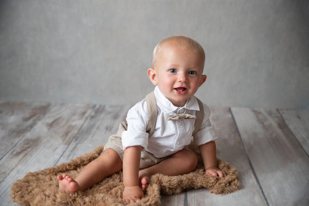 Happy baby boy in white shirt and bow tie sitting on fluffy brown rug.