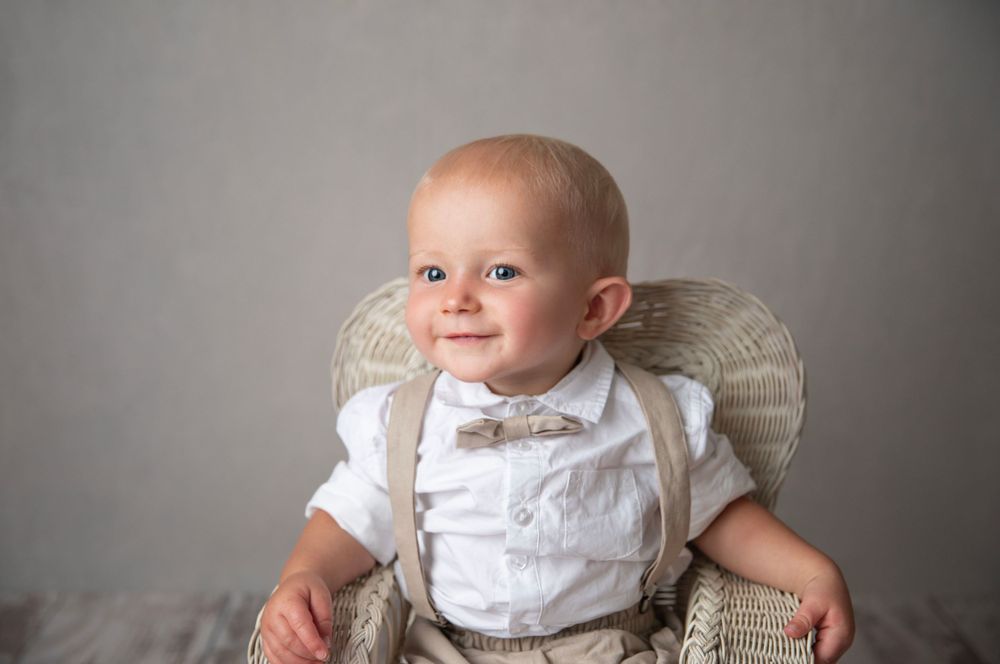Smiling baby boy in white shirt with bow tie and suspenders in wicker chair.
