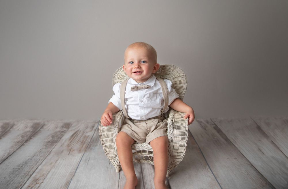 Happy baby boy in white shirt and khaki suspenders sitting in a wicker chair on wood floor.