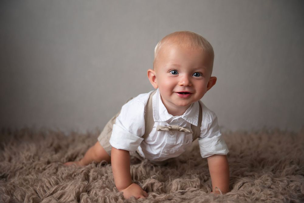 Adorable baby boy in white shirt and bow tie crawling on a fluffy beige fur rug.
