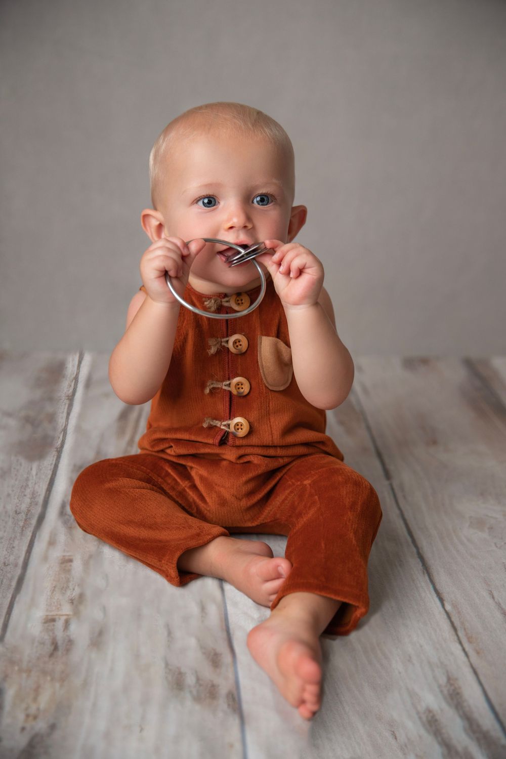 Curious baby in rust corduroy romper sitting on wood floor chewing on a silver bangle bracelet.