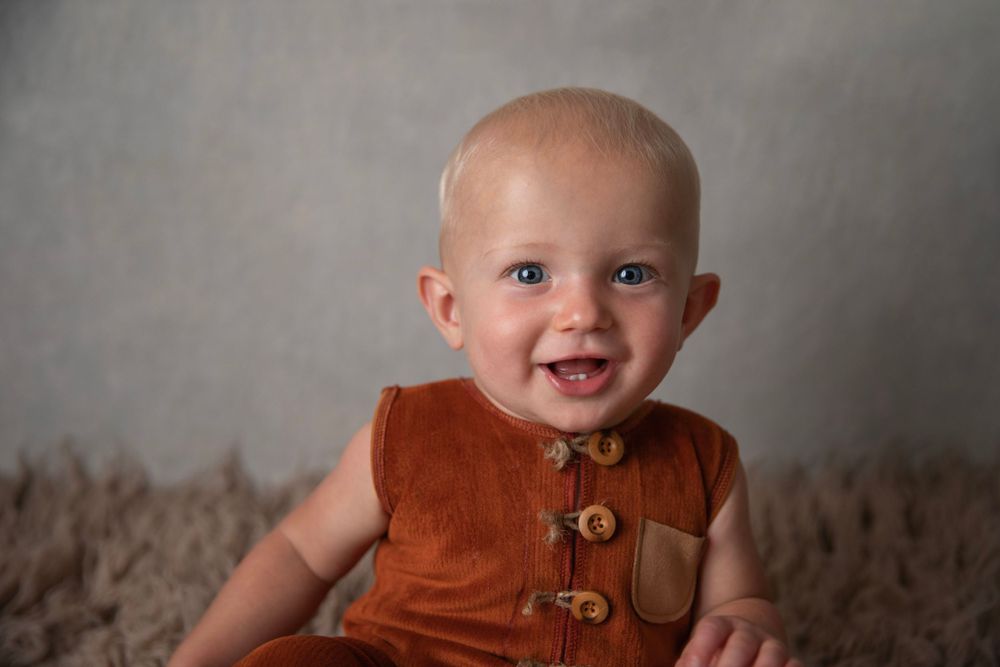 Smiling baby boy in rust brown corduroy romper sitting up against a neutral gray background.