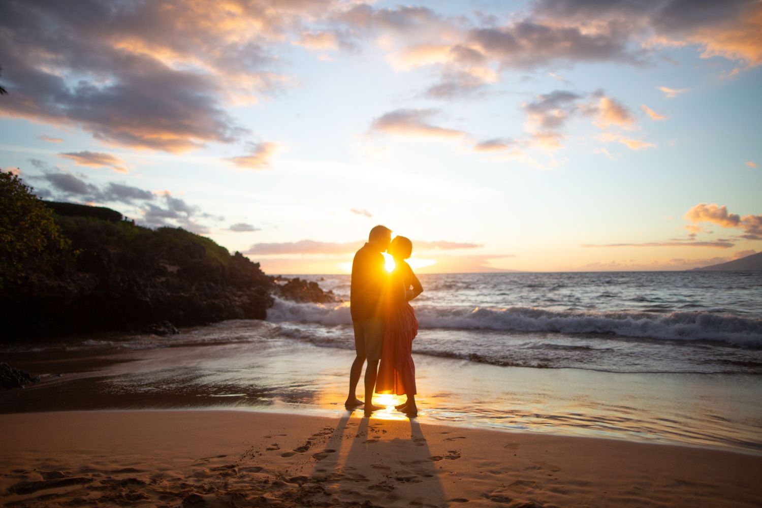 A silhouetted couple embraces on a beach during a dramatic sunset with clouds overhead.