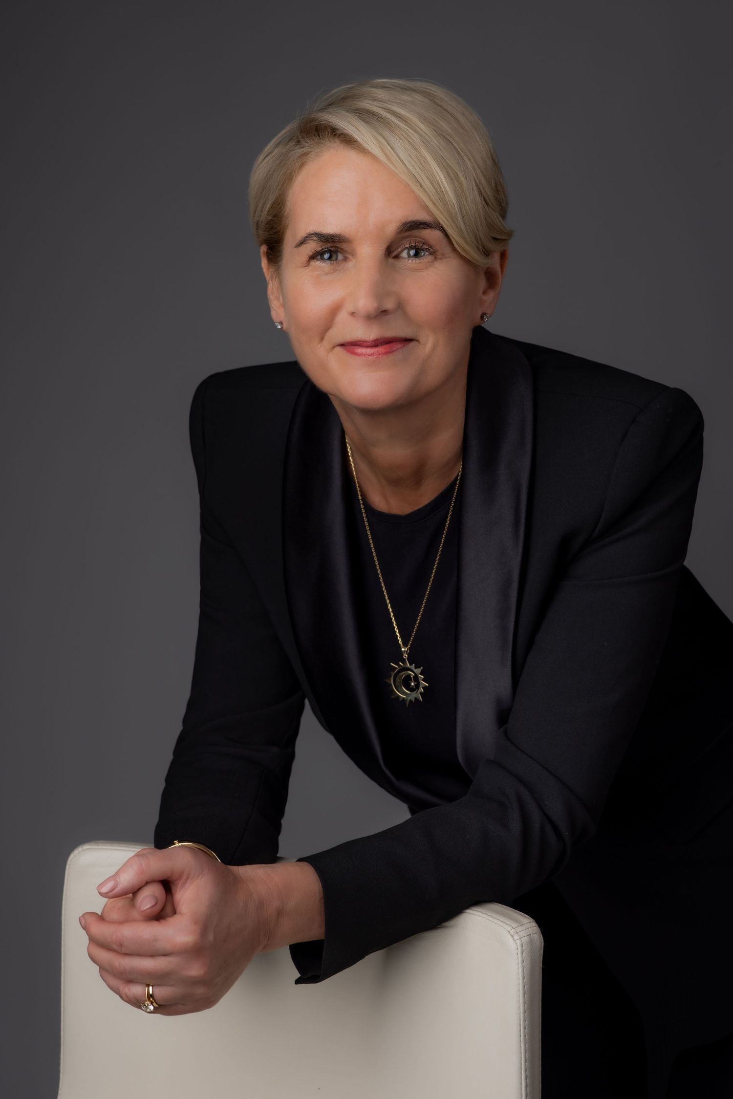 A professional portrait of a woman with short blonde hair, wearing a dark blazer and leaning on a chair, set against a studio background.