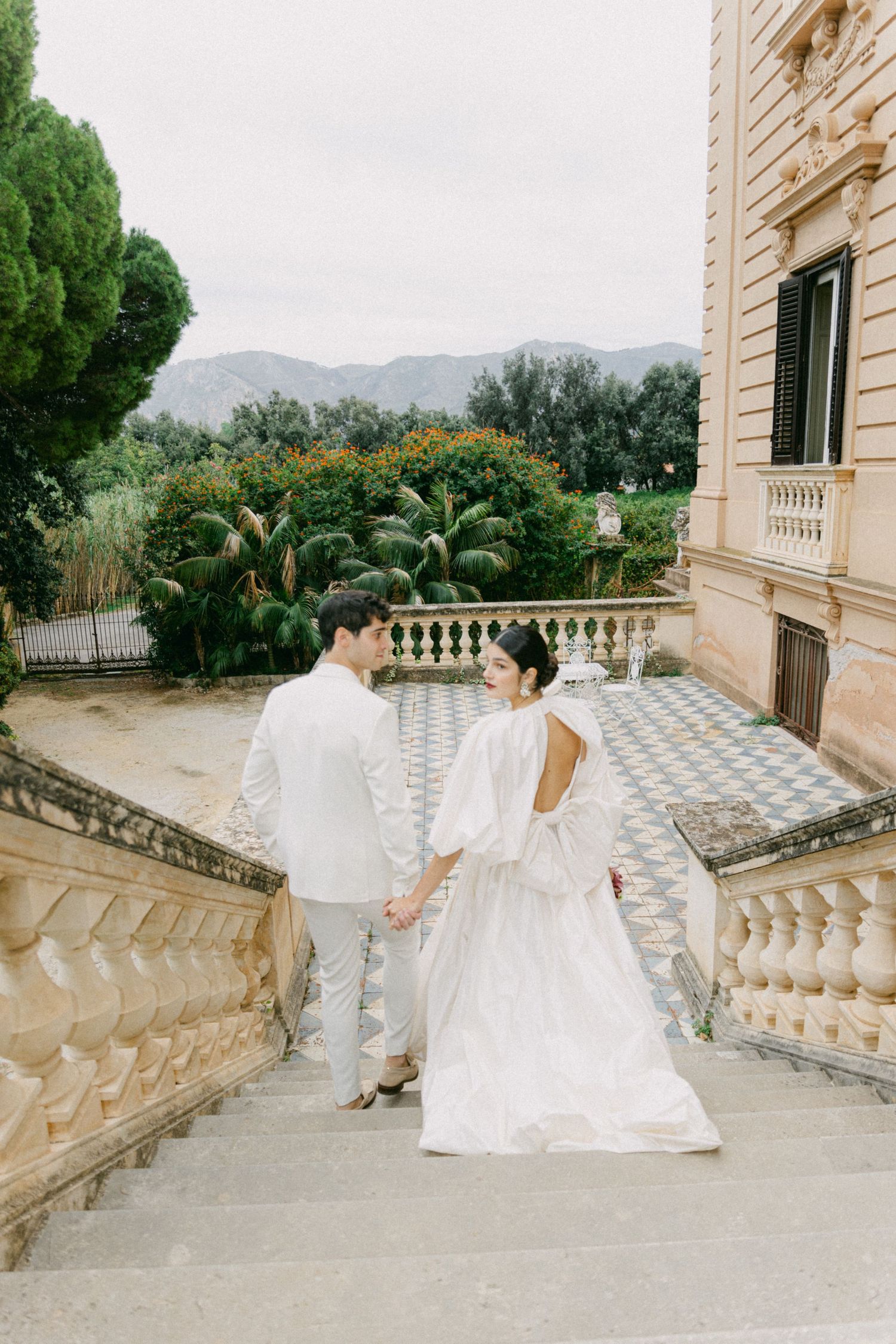 Elegant couple in white attire standing on ornate stone balcony with mountain views.