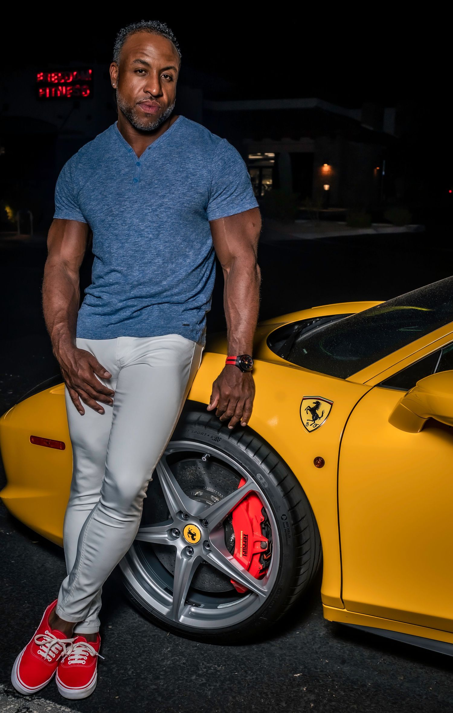 Person posing with a yellow Ferrari sports car at night, showing off the wheel and red brake caliper.