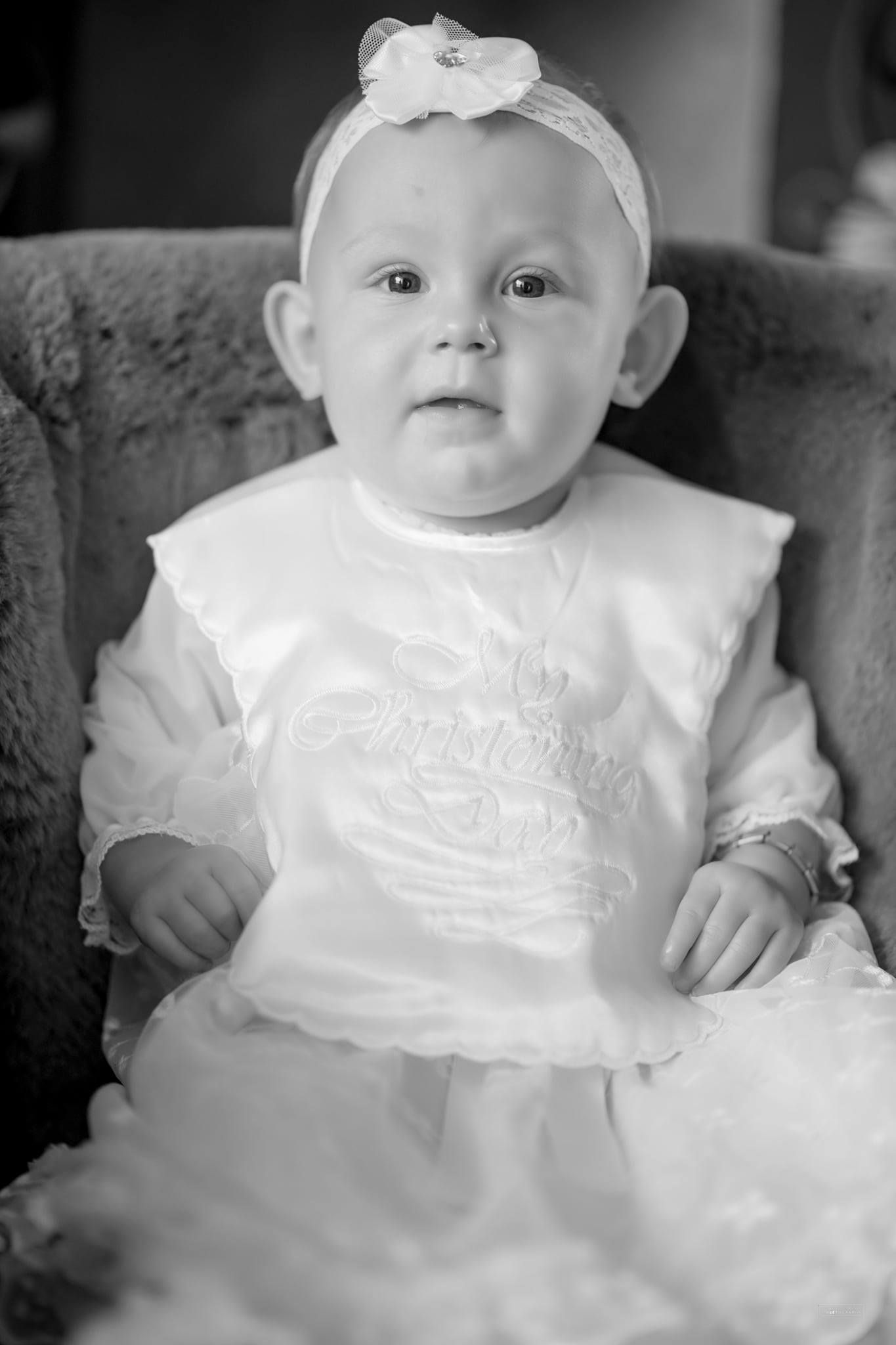 Baby in white christening gown and headband sits in vintage chair for portrait.