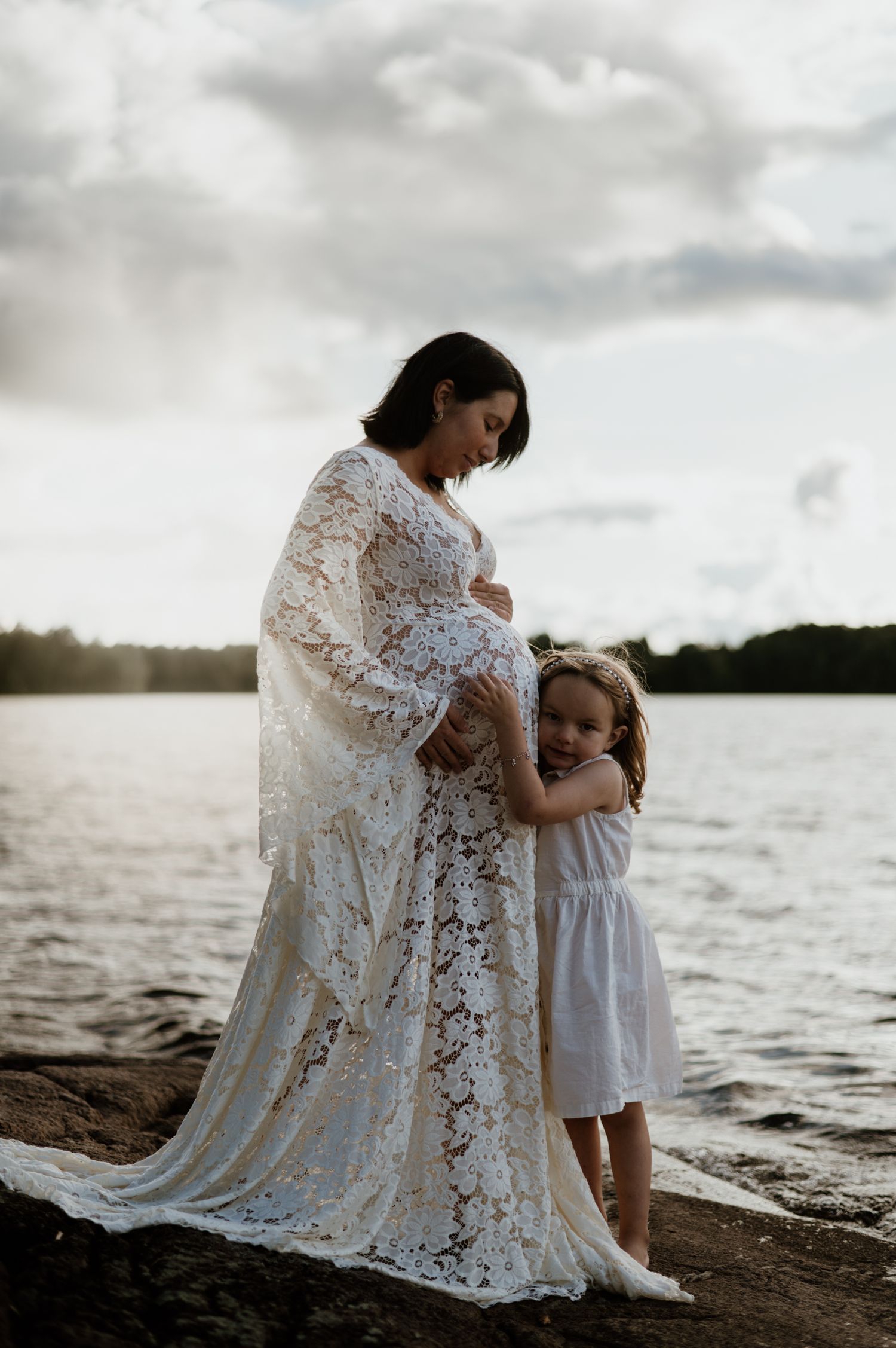 Gravid kvinna med dotter står på klipporna i solnedgång.