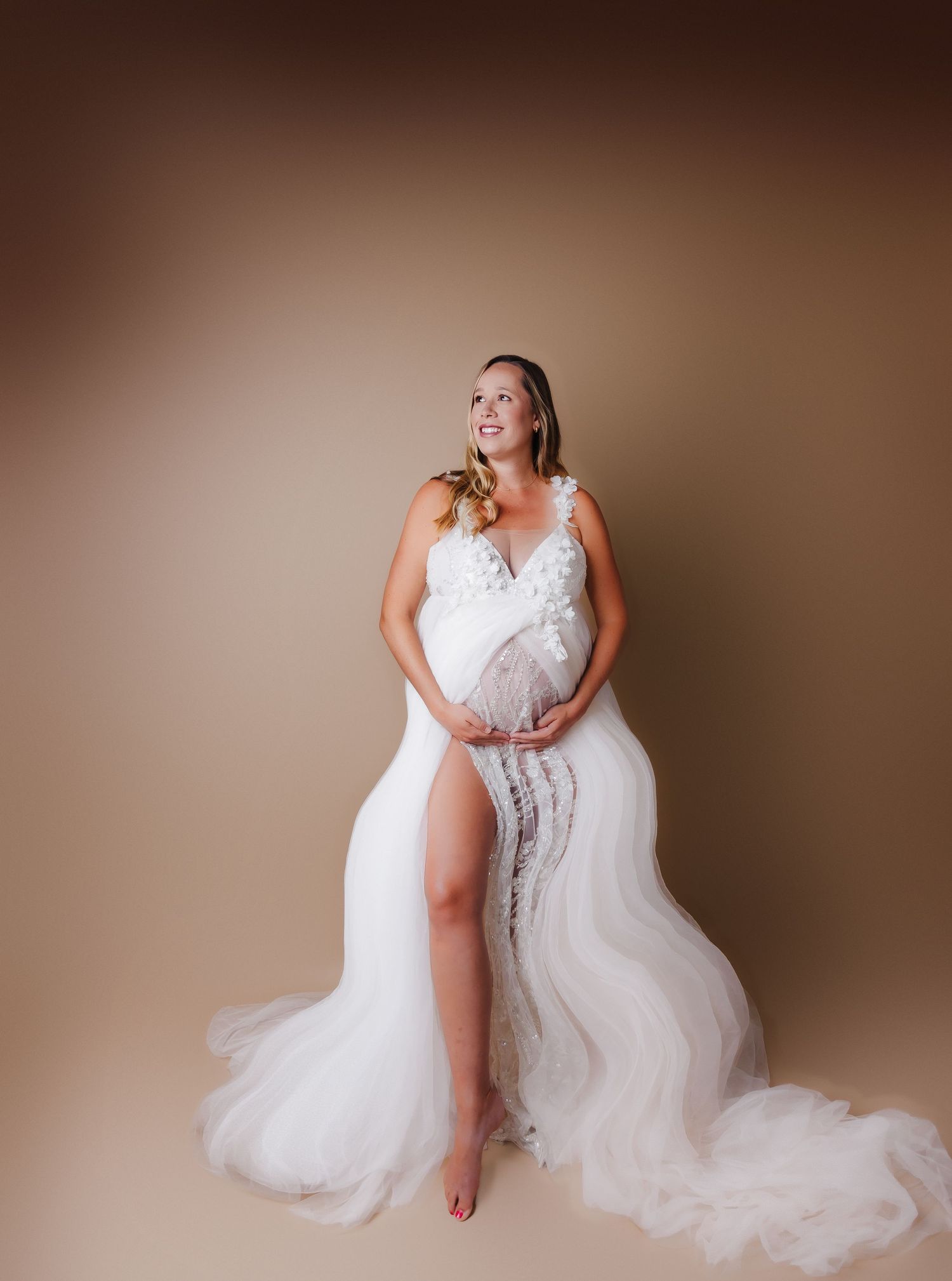 Elegant maternity photo of woman in flowing white lace gown against brown backdrop.