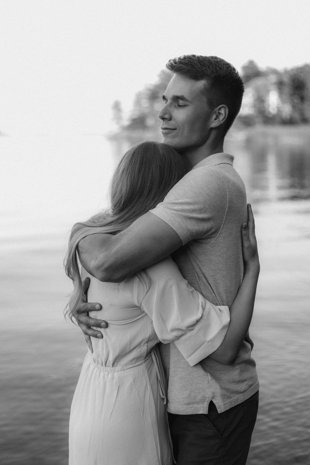 A romantic couple embracing by a serene lake in black and white photography.