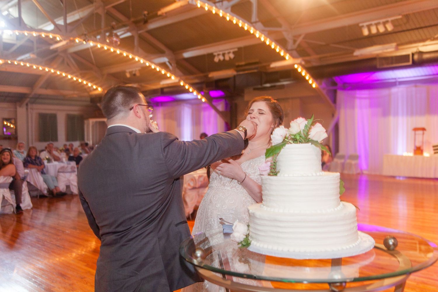 Wedding reception venue decorated with string lights and purple uplighting during cake cutting ceremony.