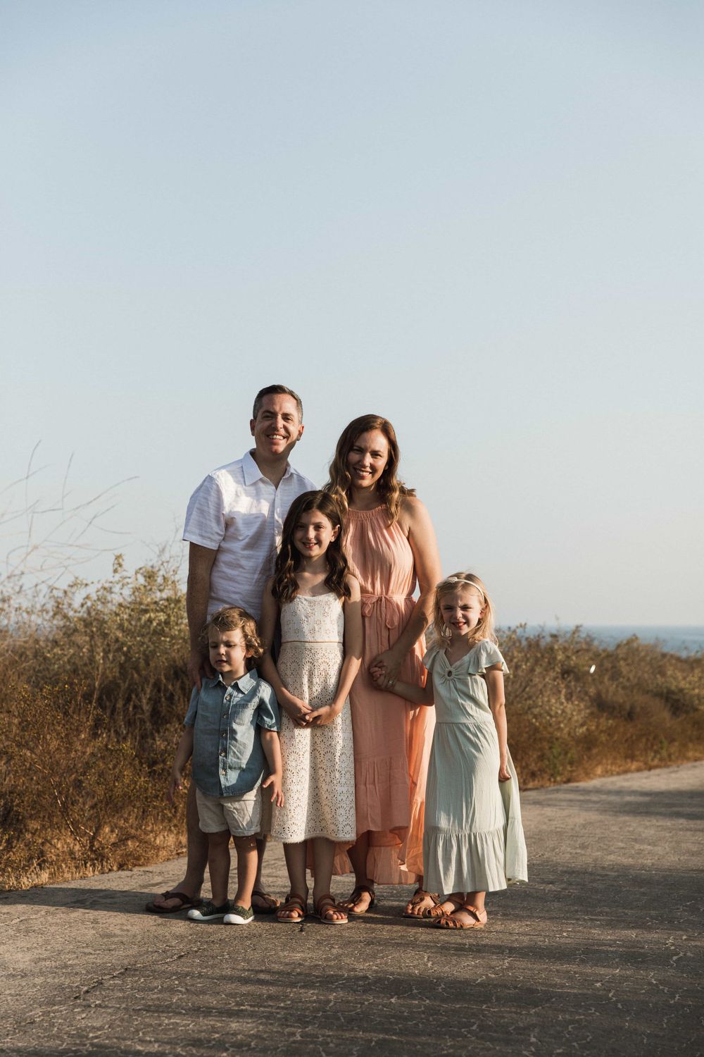 Happy family of five posing on a coastal path at golden hour with ocean and brush in background.