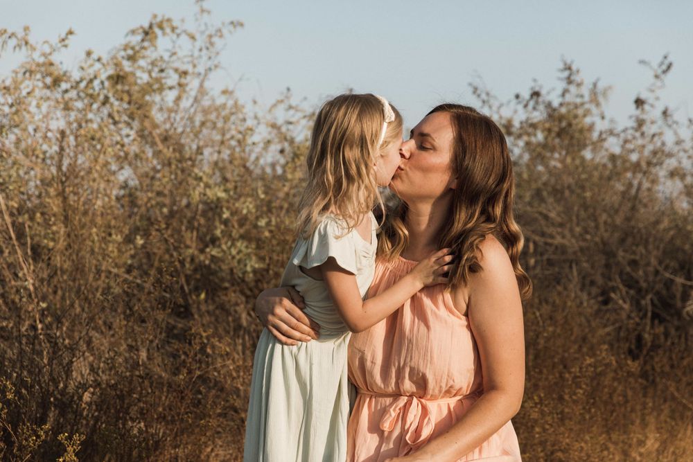 A young girl kisses her smiling mother on the cheek outdoors in warm golden sunlight.