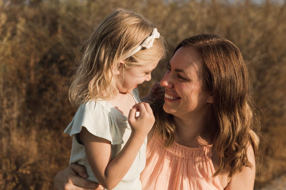 A laughing mother holds her blonde toddler daughter in a sun-drenched field of golden grass.