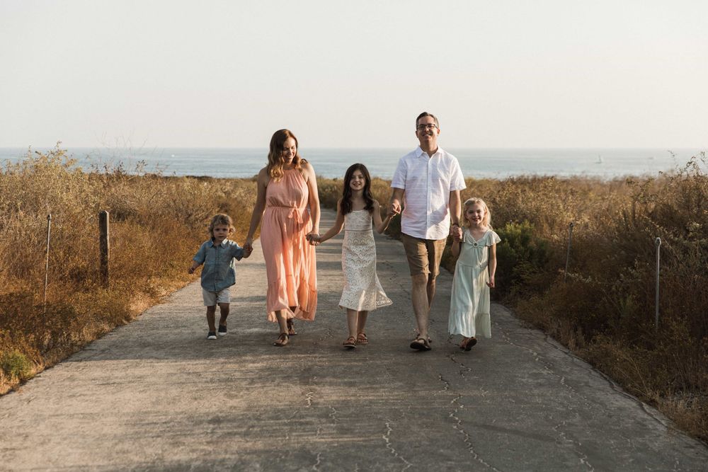 Smiling family of five strolling together on a sunny coastal pathway with ocean in background.