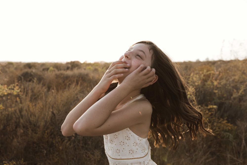 A joyful young girl laughs with hands on face in a sunlit golden field at sunset.