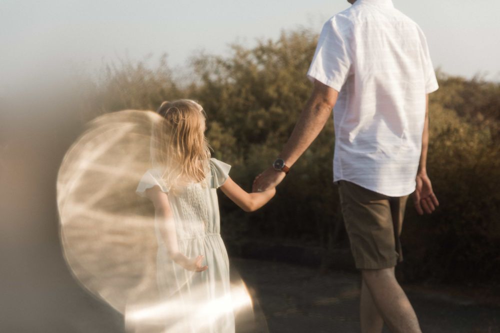 A young girl holds her fathers hand while walking along a sun-drenched outdoor path.