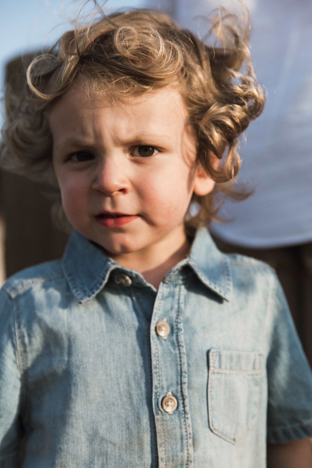 A curly-haired toddler in a denim shirt looks curiously at the camera outdoors.