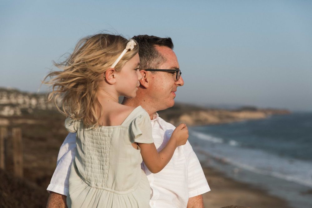 A father holds his young blonde daughter near a scenic beach overlook on a sunny day.