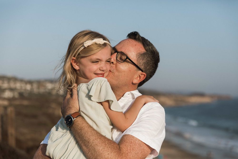 A father lovingly kisses his smiling young daughter by the beach on a clear day.
