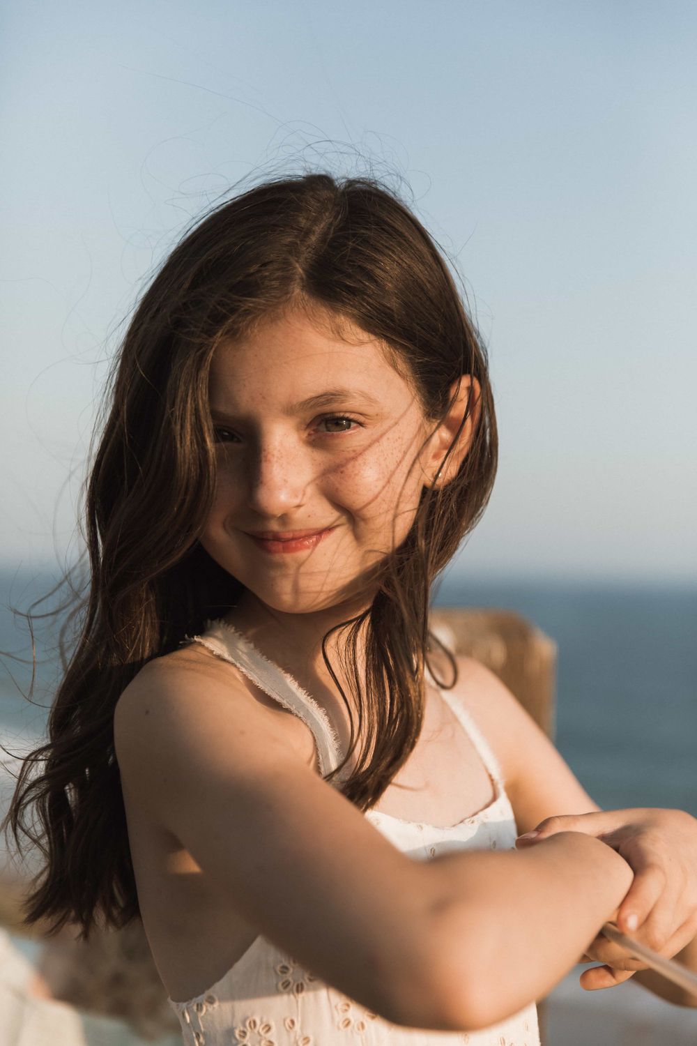 A smiling dark-haired girl in a white dress poses by the ocean on a sunny day.