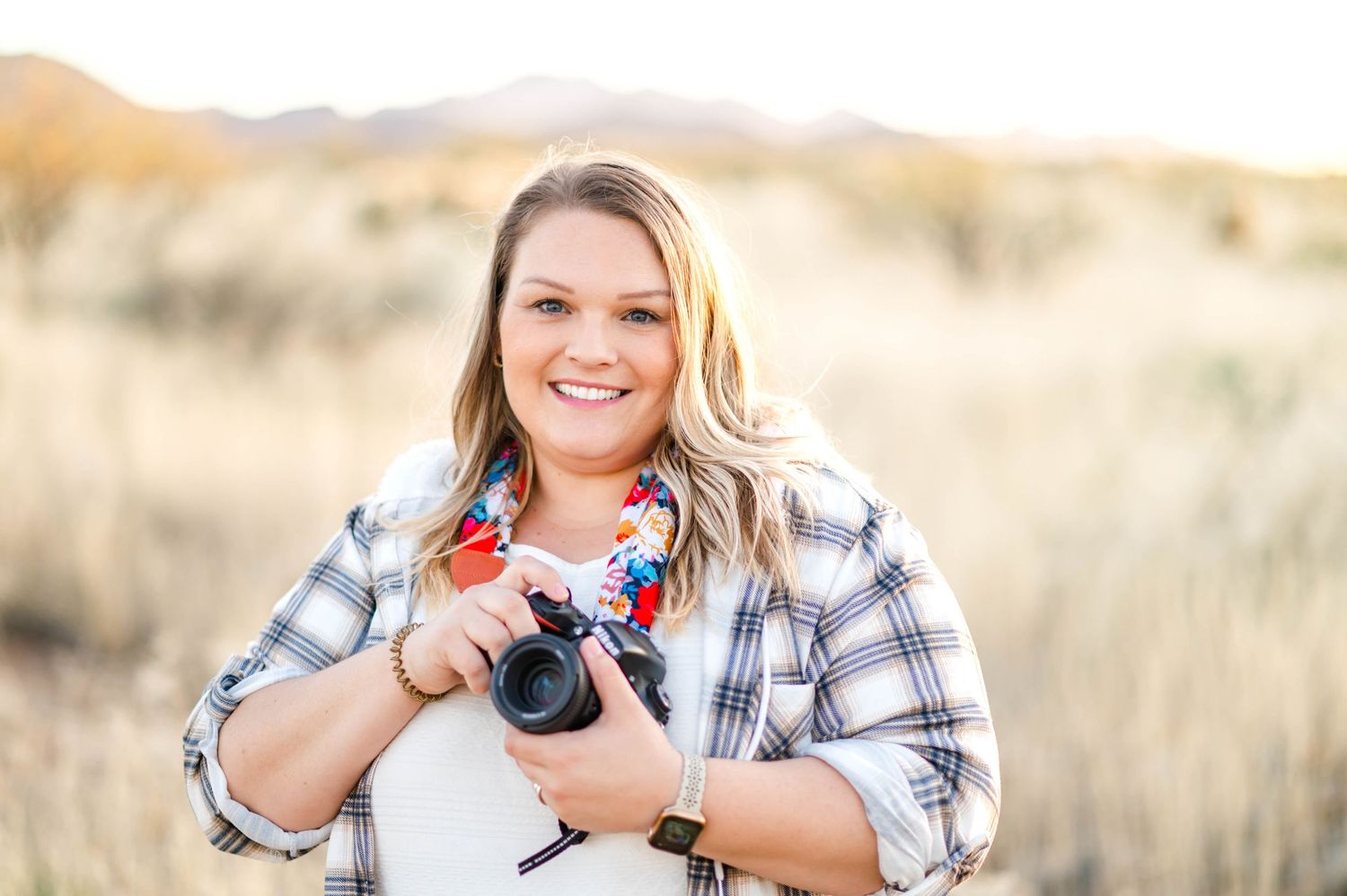 Smiling female photographer holding DSLR camera in open desert field wearing plaid shirt at golden hour.