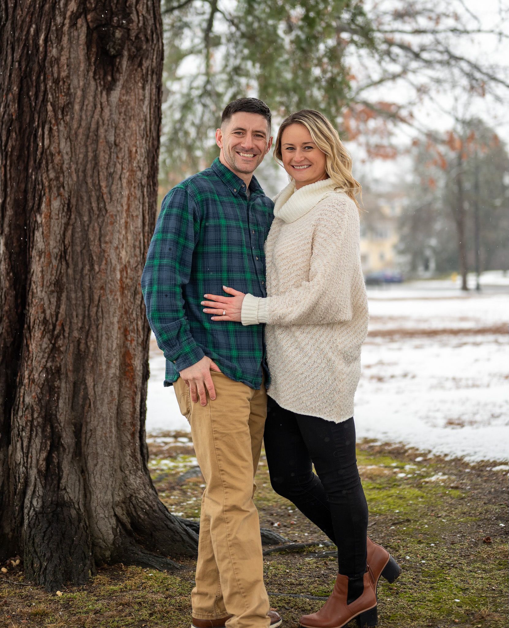 Couple posing together near large tree trunks in winter setting.