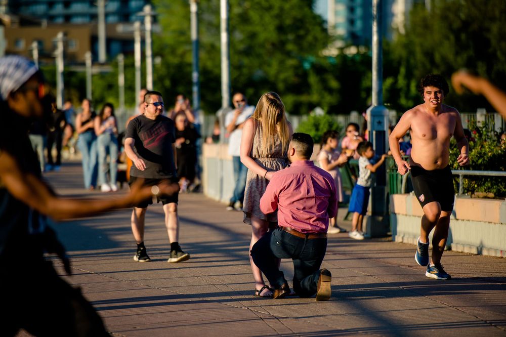 A surprise proposal moment unfolds on a crowded boardwalk with city buildings in the distance.
