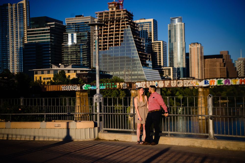 A couple stands on a bridge with the Austin skyline visible in the background on a sunny day.