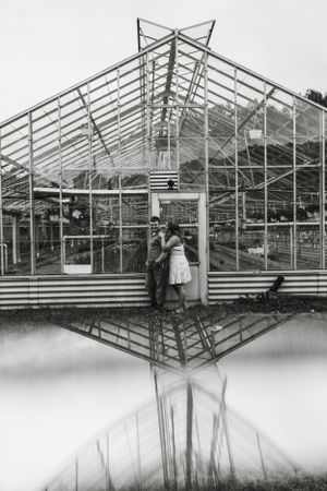A black and white photo of a couple by a greenhouse with a reflection in water.