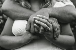 Close-up black and white photo of hands with engagement ring interlocked in embrace.
