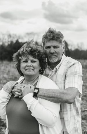Black and white portrait of mature couple embracing in casual attire against rural backdrop.
