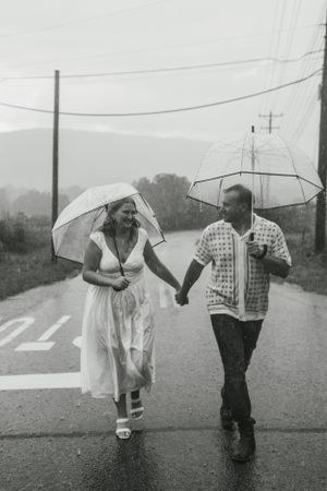 A couple laughs while walking with umbrellas in the rain on a country road.