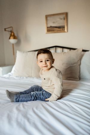 Child wearing cream sweater sitting on white bedding with wall lamp and framed art.