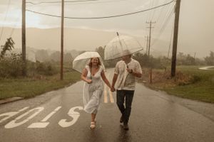A couple runs playfully in the rain holding umbrellas on a rural road.