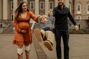Energetic family photo with child jumping while parents laugh on campus grounds.