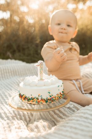 Baby in tan outfit sits next to first birthday cake with white frosting and sprinkles in warm sunlight.