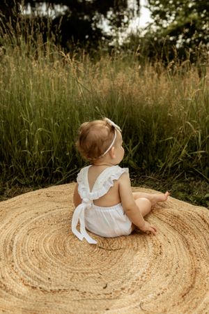 A small figure in white clothing sits on a circular jute rug surrounded by tall grass.