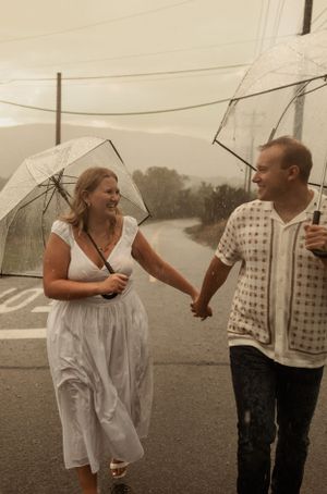 A couple walks together holding clear umbrellas during a rainy day photoshoot.